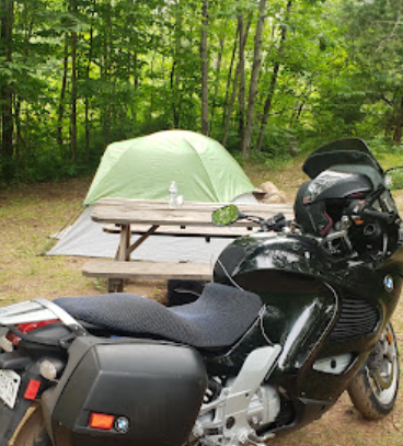 A motorcycle is parked near a picnic table with a green tent in a wooded campsite.