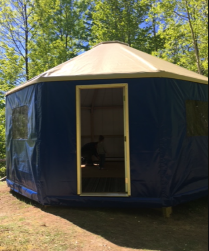 A blue yurt with a beige roof stands on a grassy area surrounded by trees. Two people are visible inside through the open door.